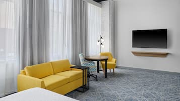 A hotel room with a yellow sofa, a round table, a TV mounted on the wall, and a patterned carpet.