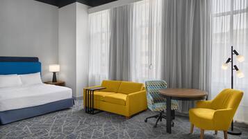 Een hotelkamer met een bed, een gele slaapbank, een blauwe fauteuil, een kleine ronde tafel en een vloerlamp.