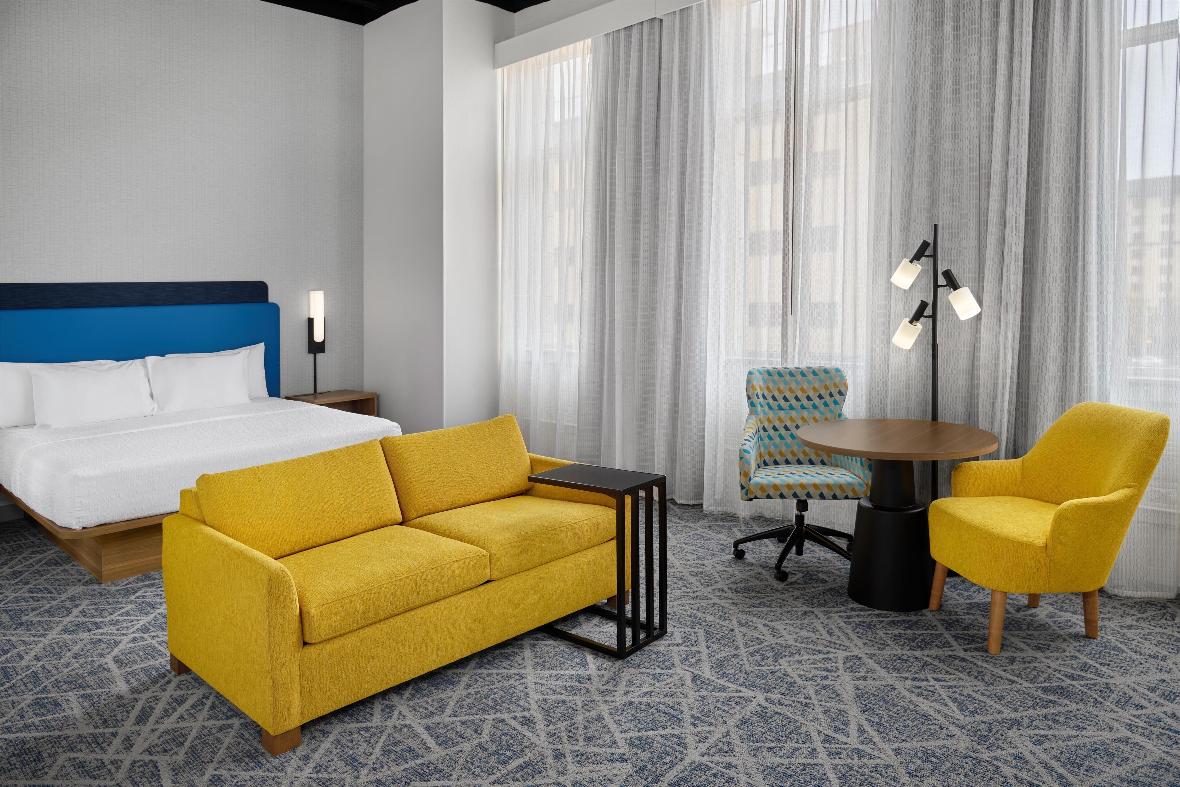 A hotel room with a bed, a yellow sofa, a small table, a chair, and a floor lamp.