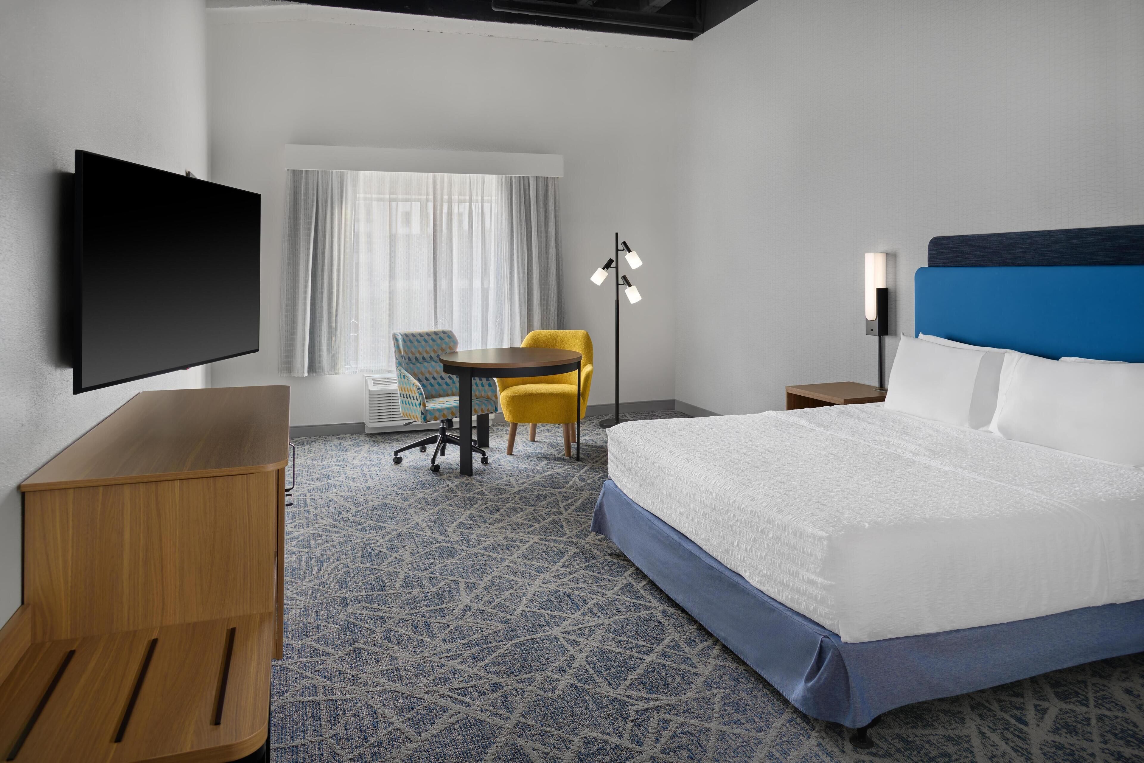 A hotel room with a large bed, a desk, a chair, a TV, and a window with curtains.