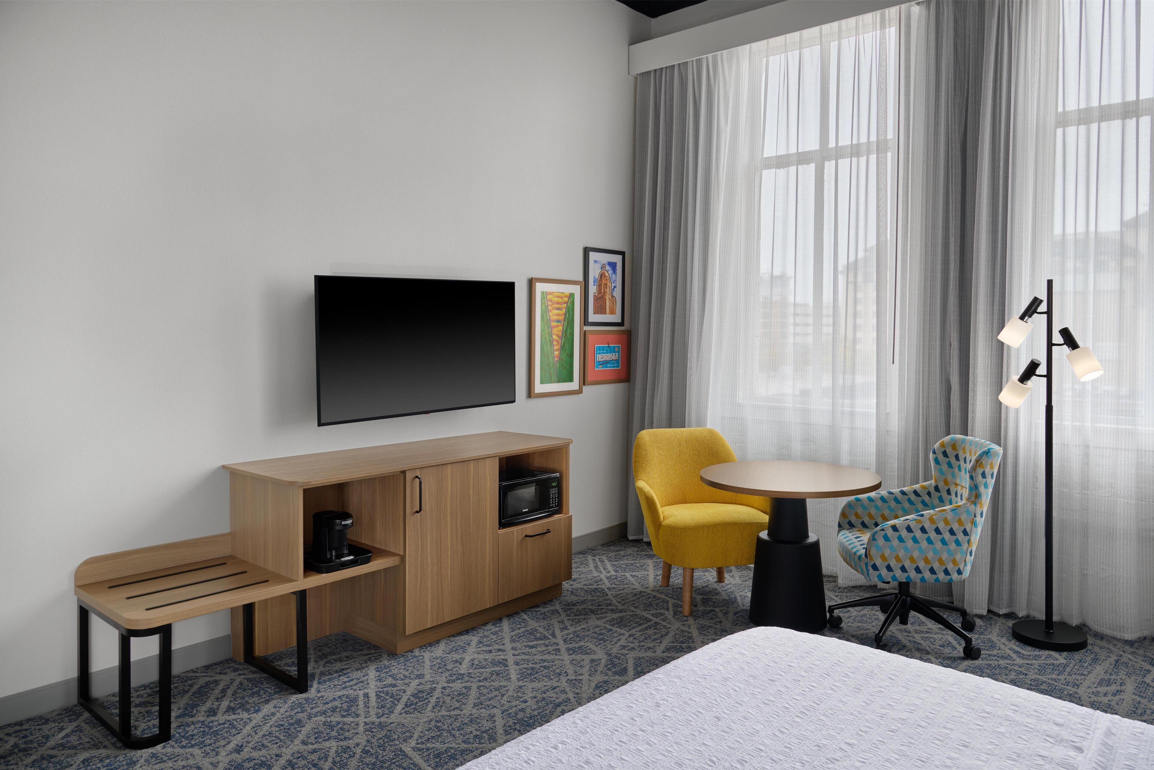 A hotel room with a bed, a TV, a wooden cabinet with a microwave, a small table, two chairs, and a floor lamp.