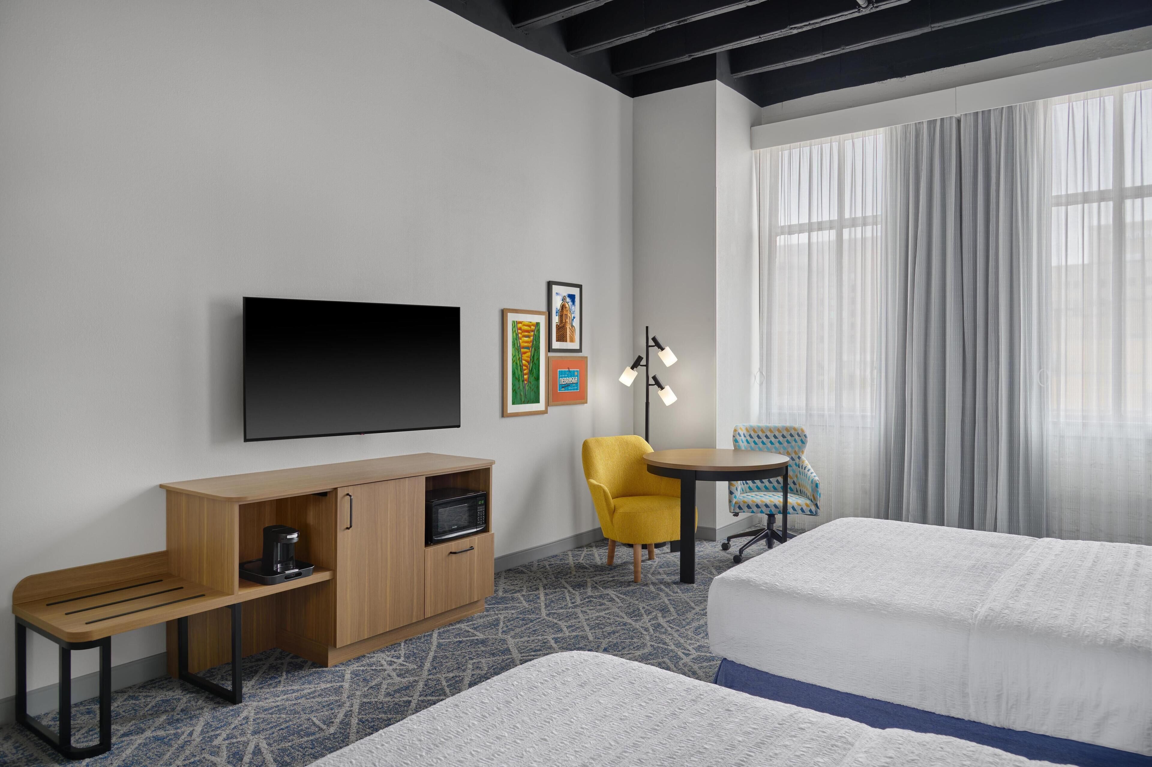 A hotel room with a bed, a desk with a microwave, a TV, a chair, and a window with curtains.