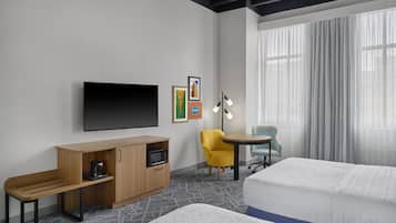 A hotel room with a bed, a desk with a microwave, a TV, a chair, and a window with curtains.