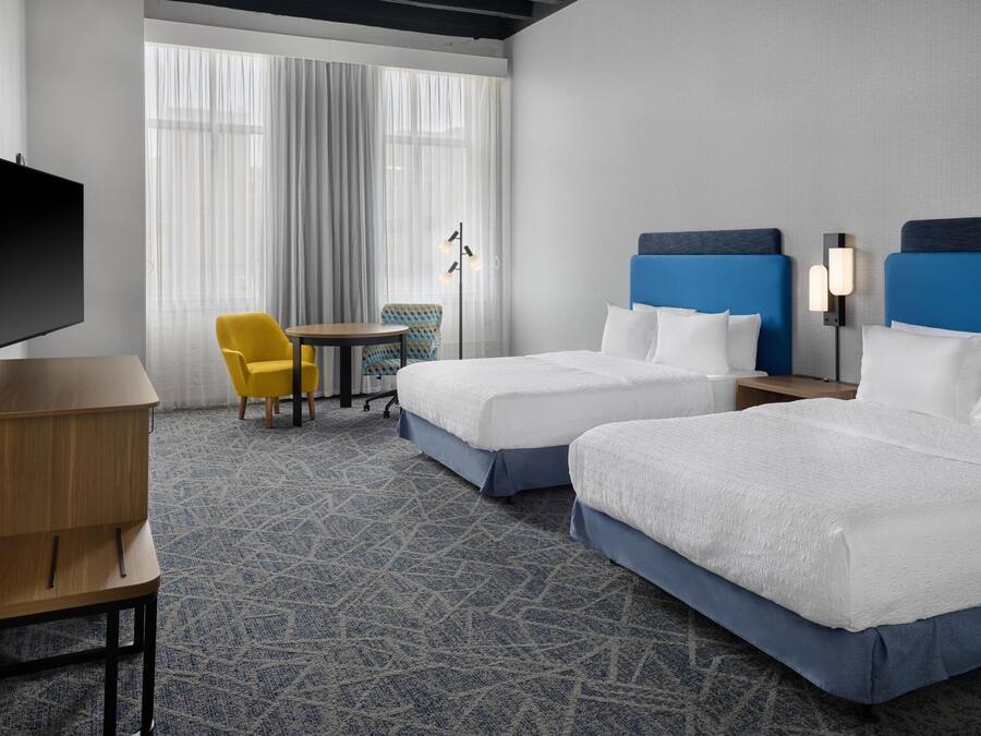 A hotel room with two beds, a desk, a chair, a TV, and a large window with curtains.