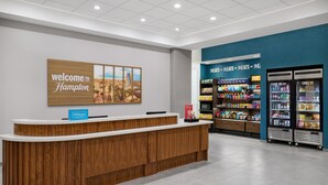 A hotel reception area with a wooden reception desk, a 'Welcome to Hampton' sign, a Hilton sign, and a snack vending area. - Hampton Inn by Hilton Lincoln Downtown (Lincoln)