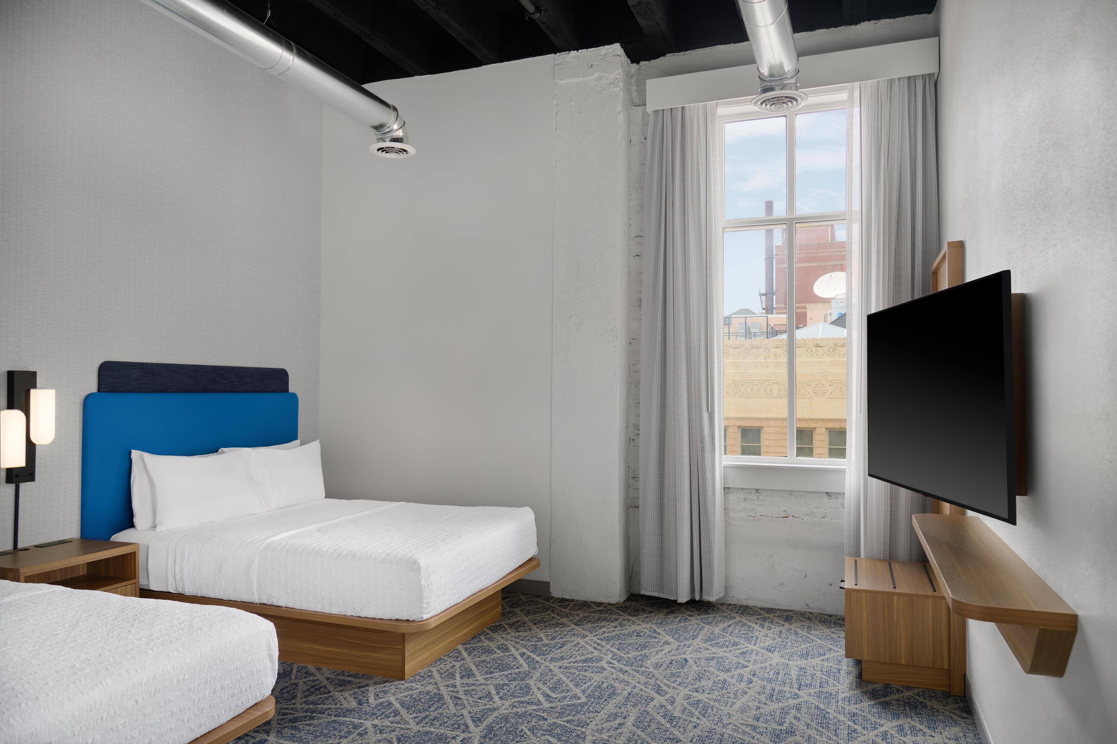 A hotel room with a bed, a TV, a window with curtains, and a patterned floor.