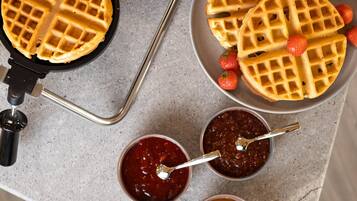 A waffle iron, waffles, strawberries, and two jam containers on a countertop.