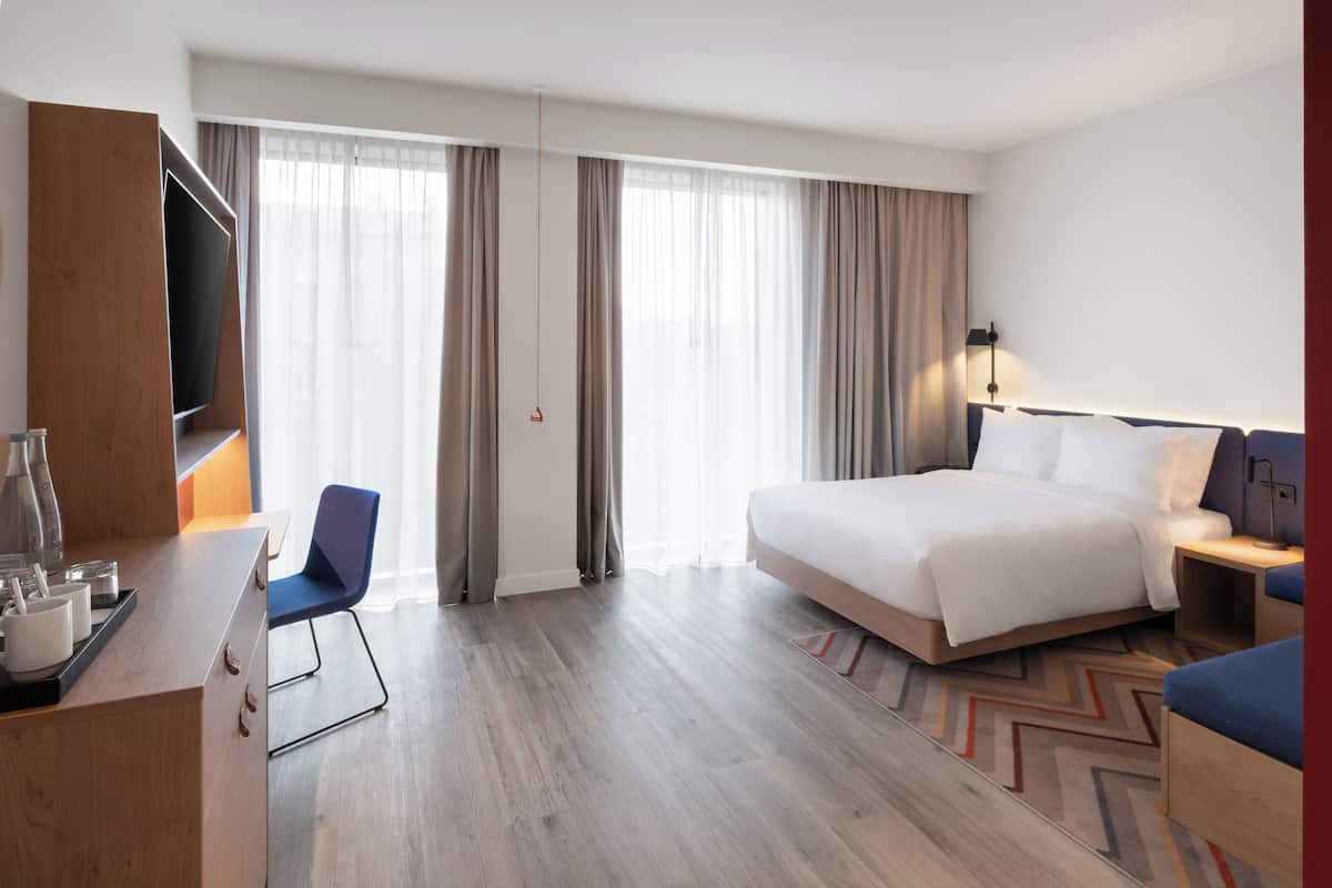 A hotel room with a bed, a desk with a chair, a TV, and a large window with curtains.
