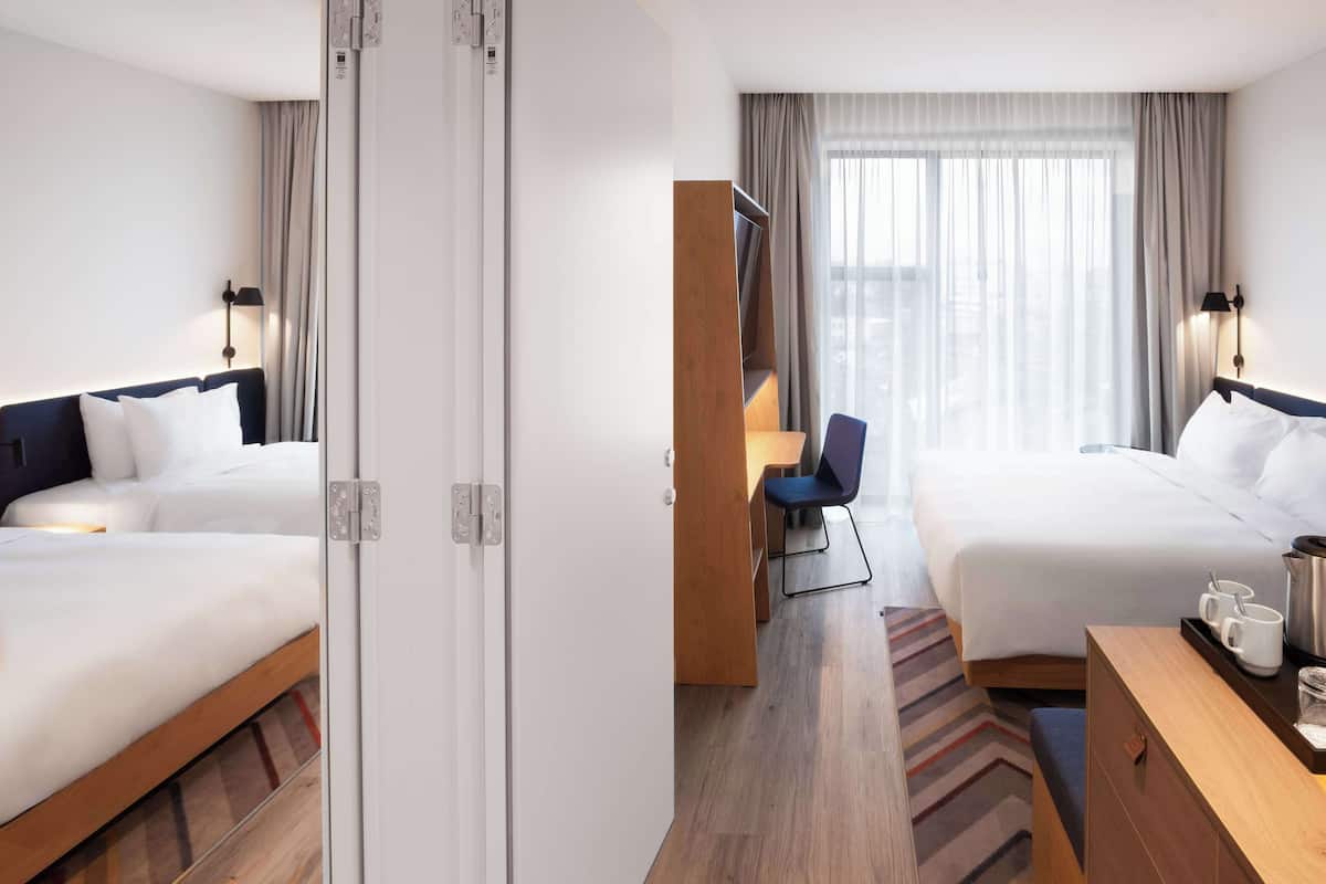 A hotel room with two beds, a desk with a tray of cups, and a view of the city through large windows.