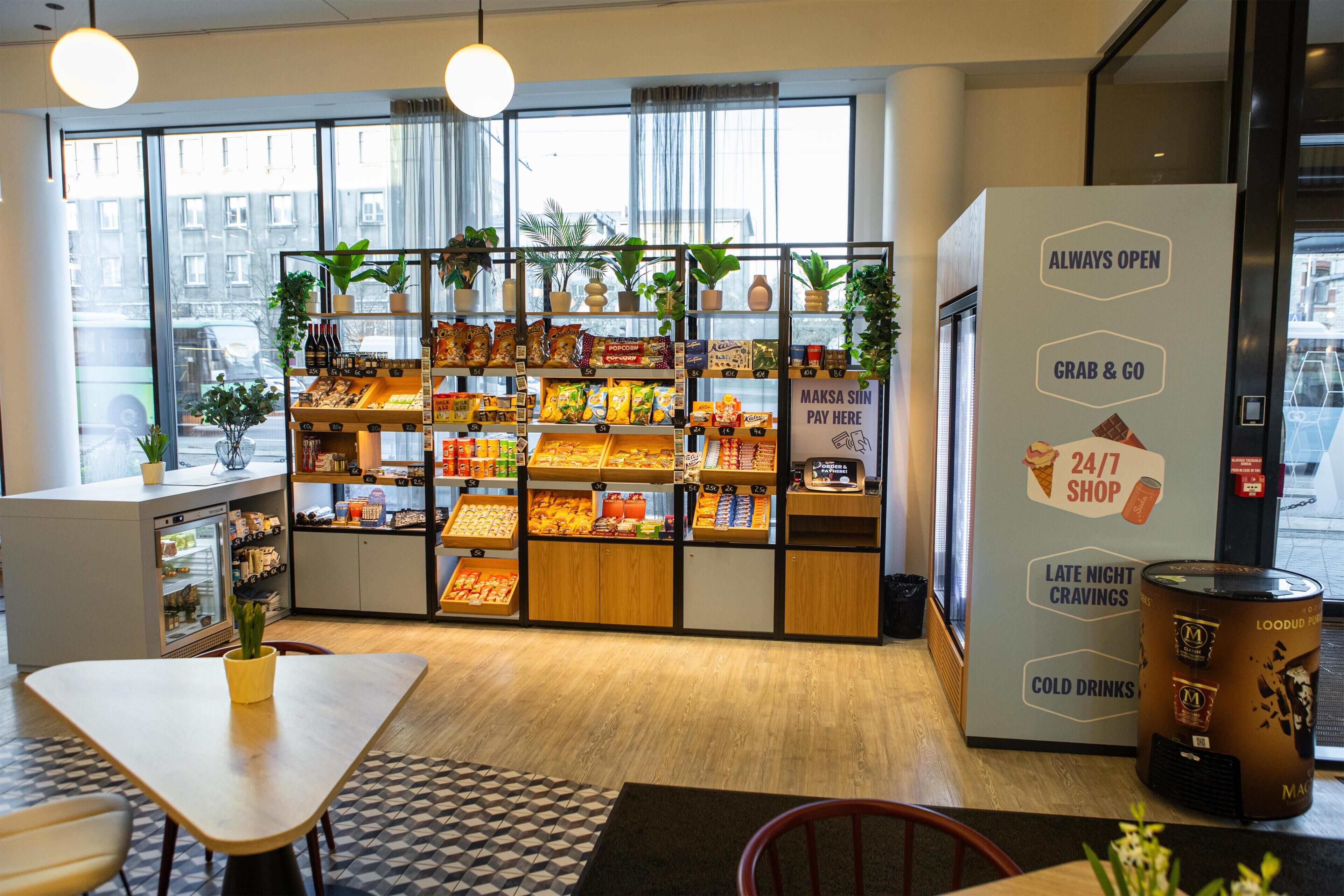 Um bar de lanches com prateleiras repletas de diversos snacks e bebidas, um caixa registradora e uma placa indicando que está sempre aberto.