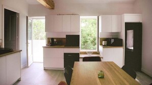 A modern kitchen with white cabinets, a wooden dining table, and a view of greenery outside. - Ahouu - Gîte with swimming pool - Pic Saint-Loup (Sauteyrargues)