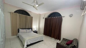 A bedroom with a bed, a nightstand, a sofa, a ceiling fan, and two windows with curtains. - Ciatle Suites & Resort  (Dhaka)