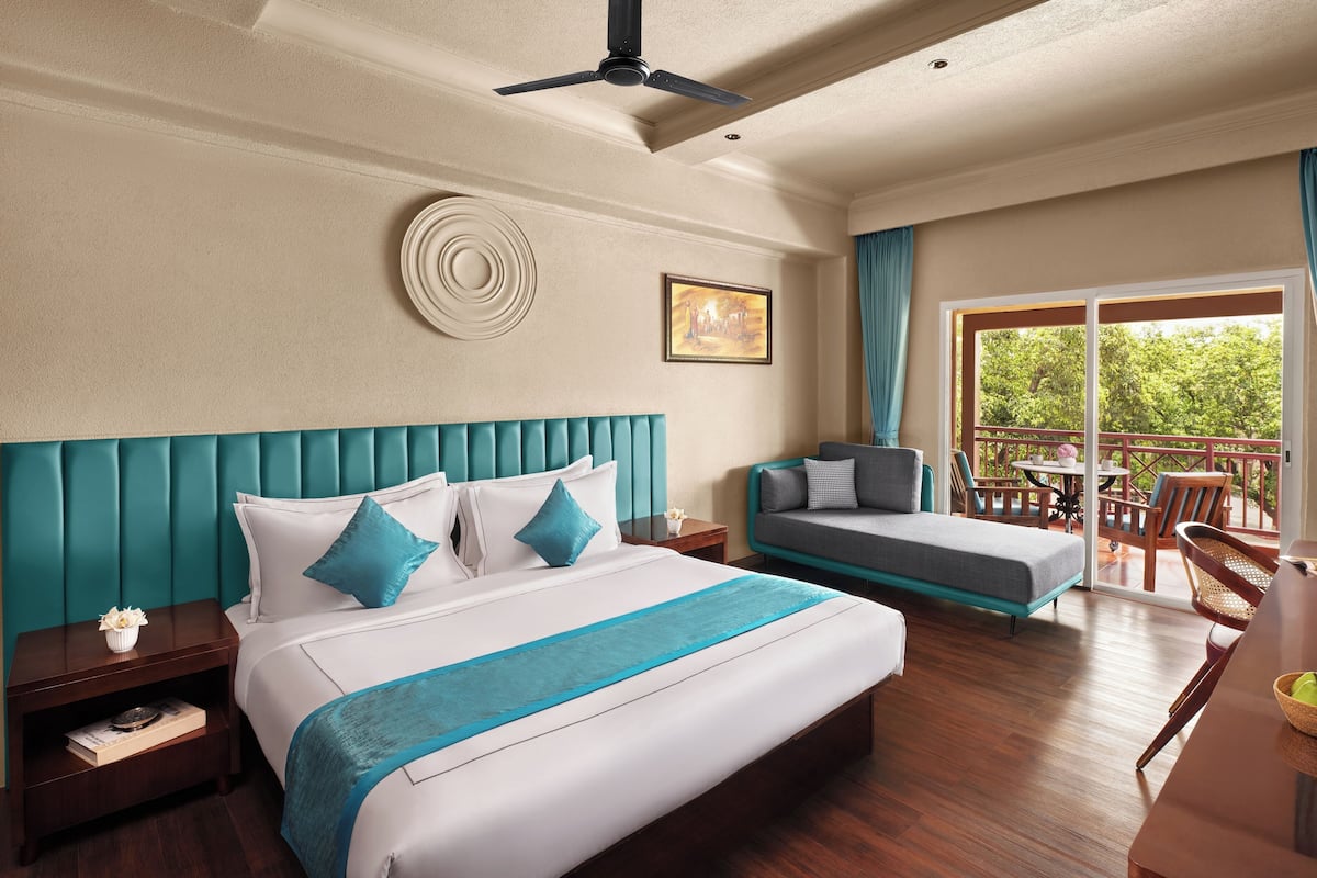 Habitación de hotel con una cama grande, un sofá, zona de comedor con sillas y un balcón con vistas a árboles.