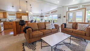 A spacious living room with a brown leather sofa, a white marble coffee table, and a black and white striped rug. There is a kitchen area with wooden cabinets and a large window offering a view of the outdoors. - Ski Winter Park/ Granby Ranch-Hot Tub- Pets-Sled- Multi-Family House (Granby)