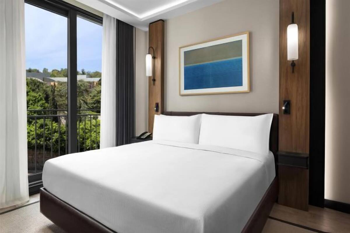 A hotel room with a large bed, a painting on the wall, and a view of greenery outside.