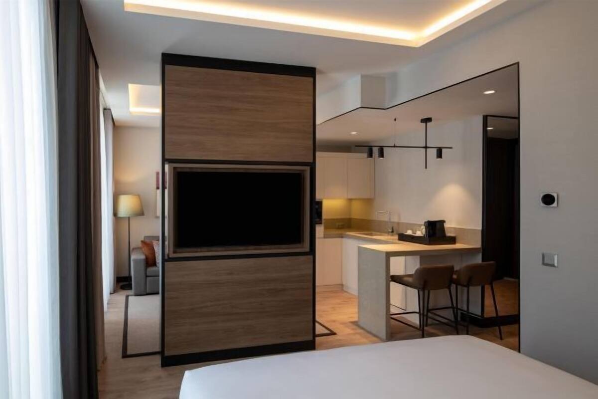 A modern hotel room with a large bed, a TV, a kitchenette, and a seating area.
