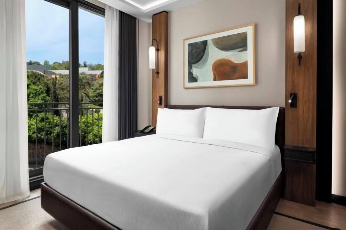 A hotel room with a large bed, a painting on the wall, and a balcony with a view.