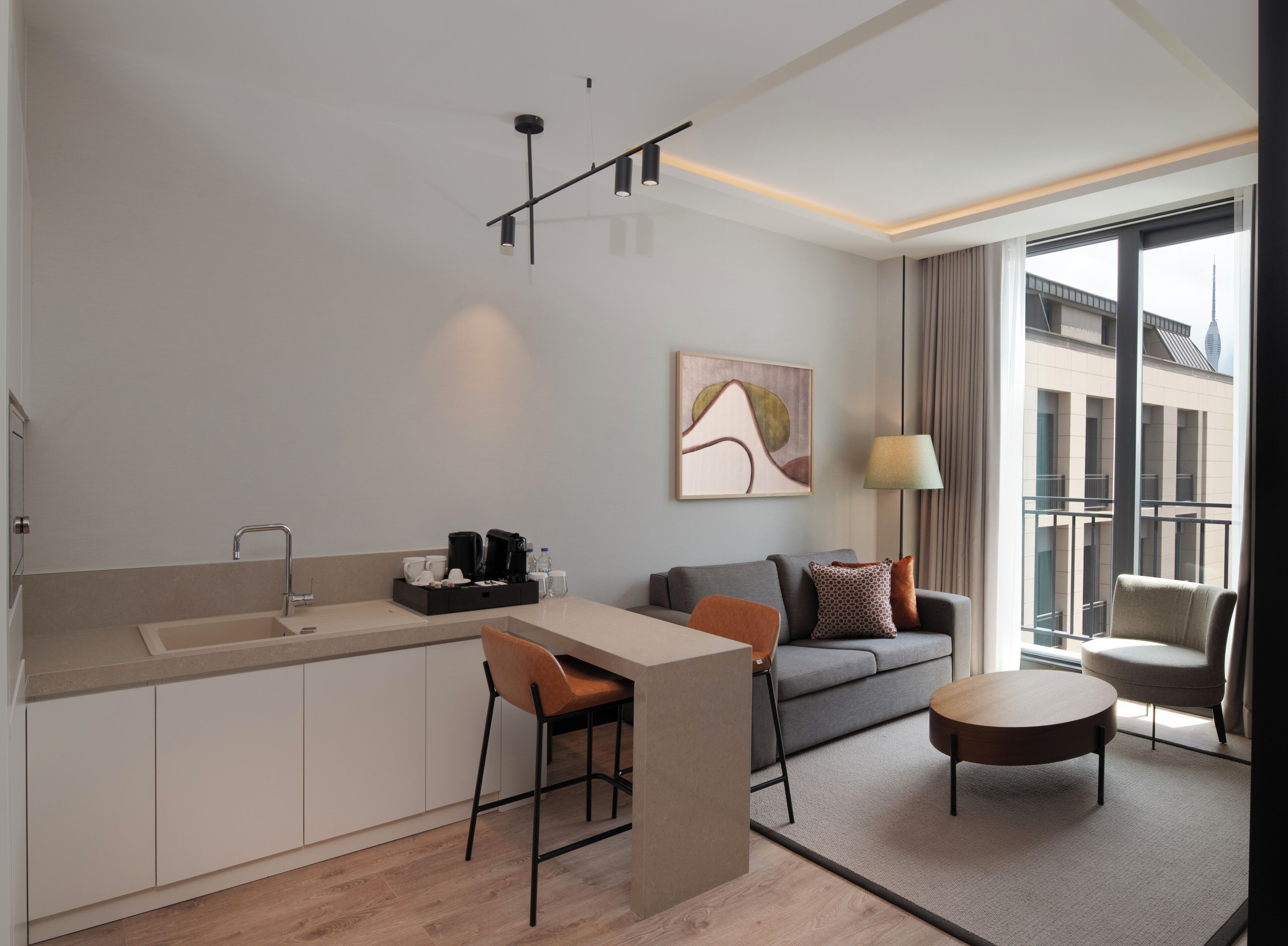 A modern kitchen with a sink, bar stools, and a coffee station. There is a sofa and a round table with chairs in the living area, and a large window with a view of buildings.