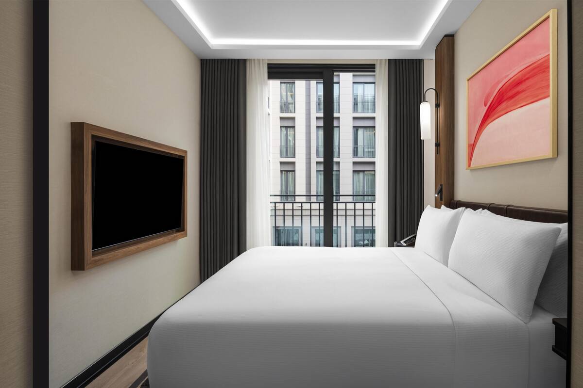 A hotel room with a large bed, a flat-screen TV mounted on the wall, a window with a view of buildings, and a modern abstract painting on the wall.