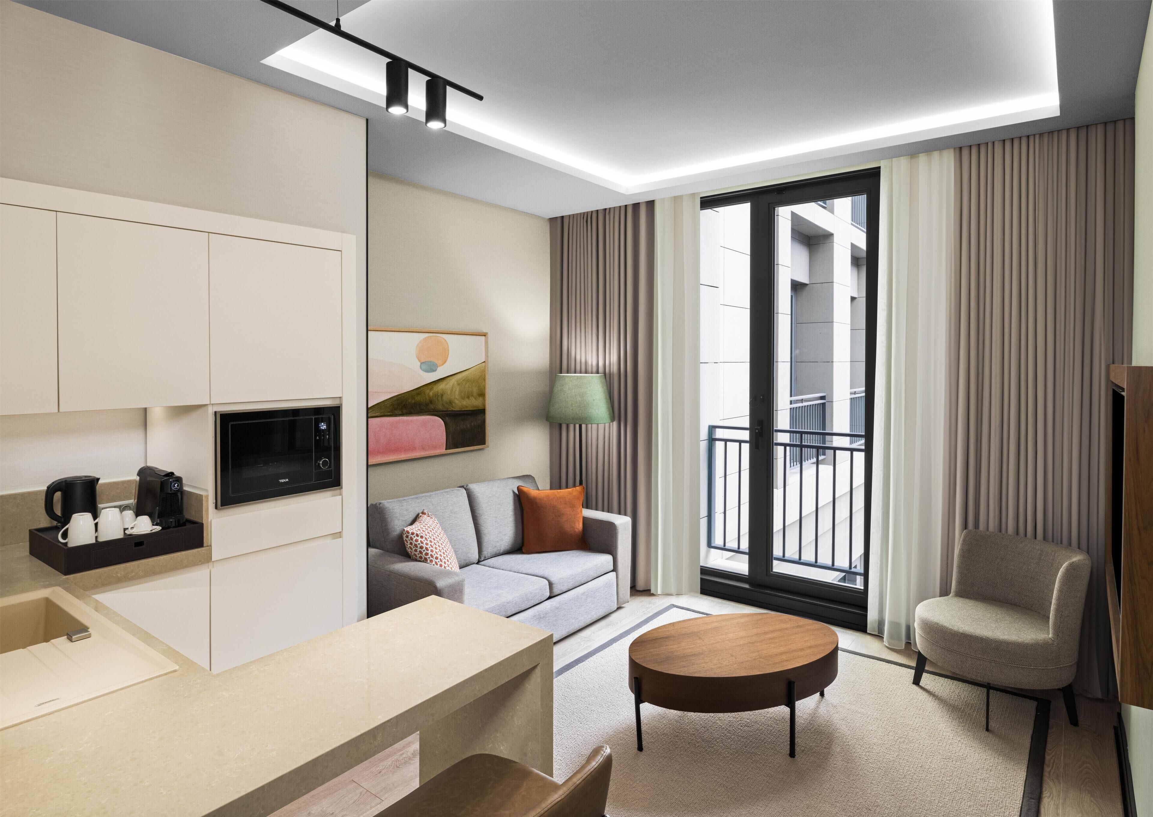 A modern hotel room with a kitchenette, a sofa, a round coffee table, a chair, and a view of the building's exterior through large windows.