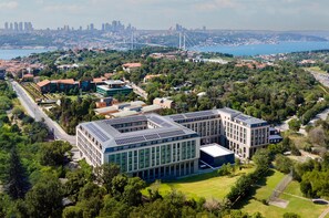 An aerial view of a modern building complex surrounded by greenery, with a city skyline and a bridge in the background. - Altunizade Suites Istanbul, Curio Collection by Hilton (Istanbul)