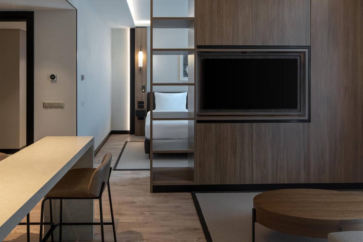 A modern hotel room with a flat-screen TV mounted on a wooden panel wall, a dining area with a table and chairs, and a separate sleeping area with a bed.