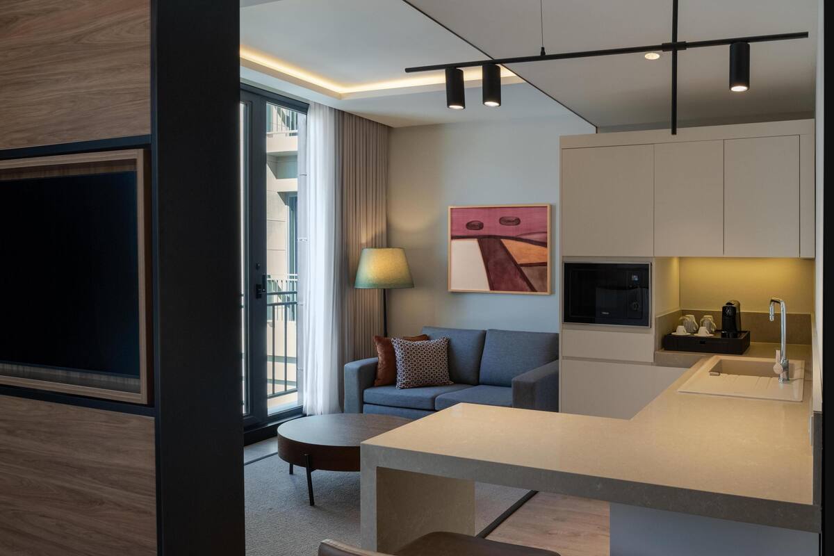 A modern hotel room with a sofa, a dining area, a kitchen with a sink, and a TV mounted on the wall.