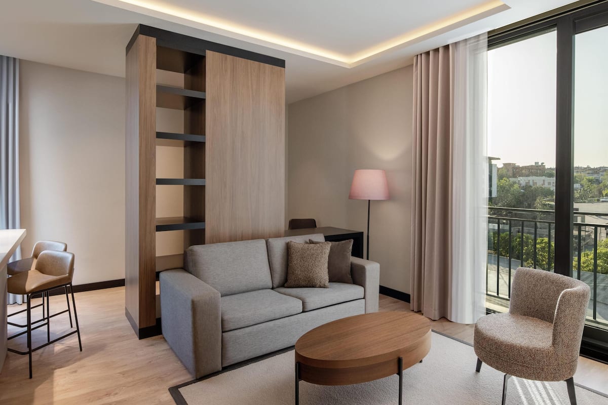 A modern hotel room with a sofa, a small table, a chair, a lamp, and a large window with a city view.