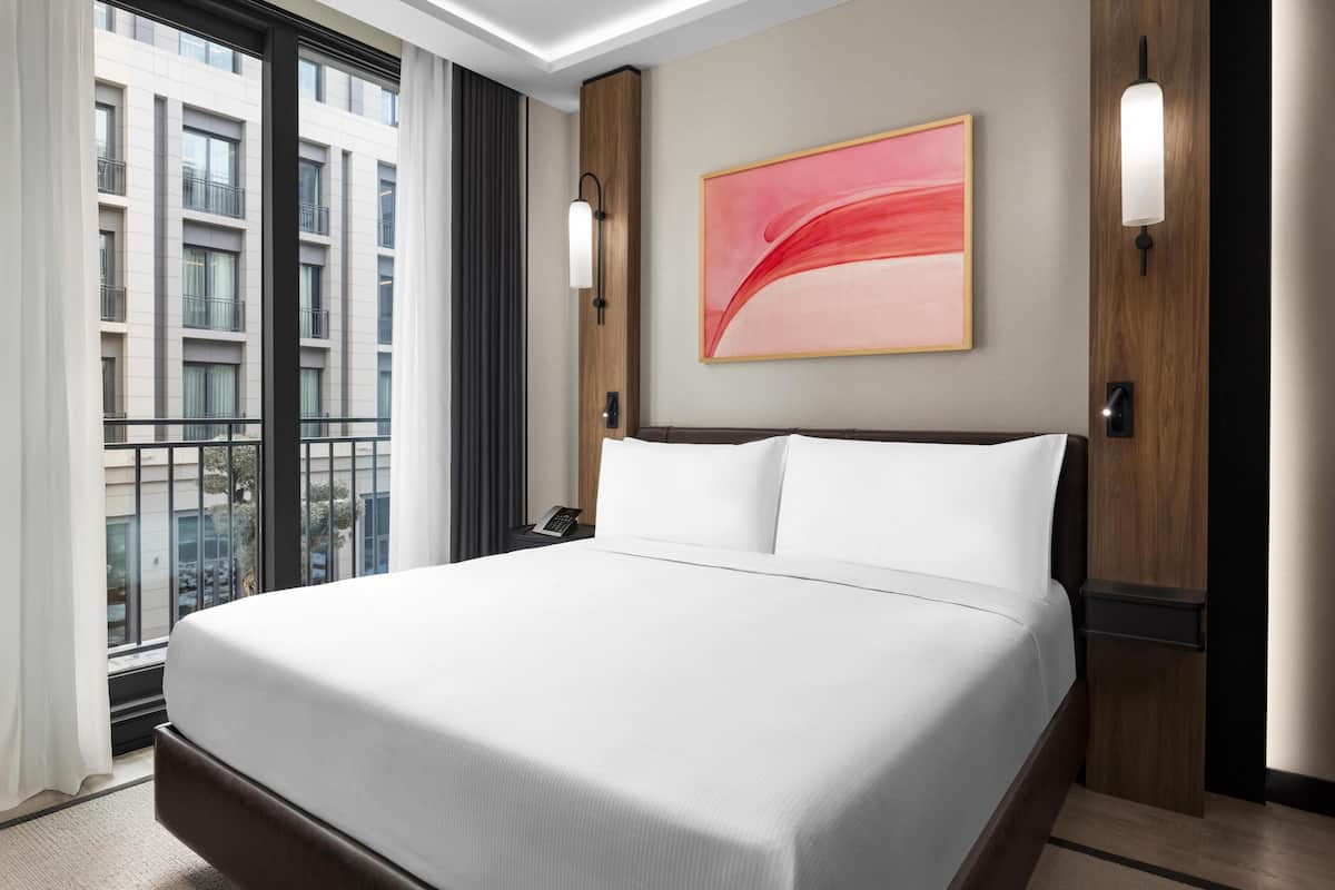 A hotel room with a large bed, a view of a building, and two wall-mounted lights.