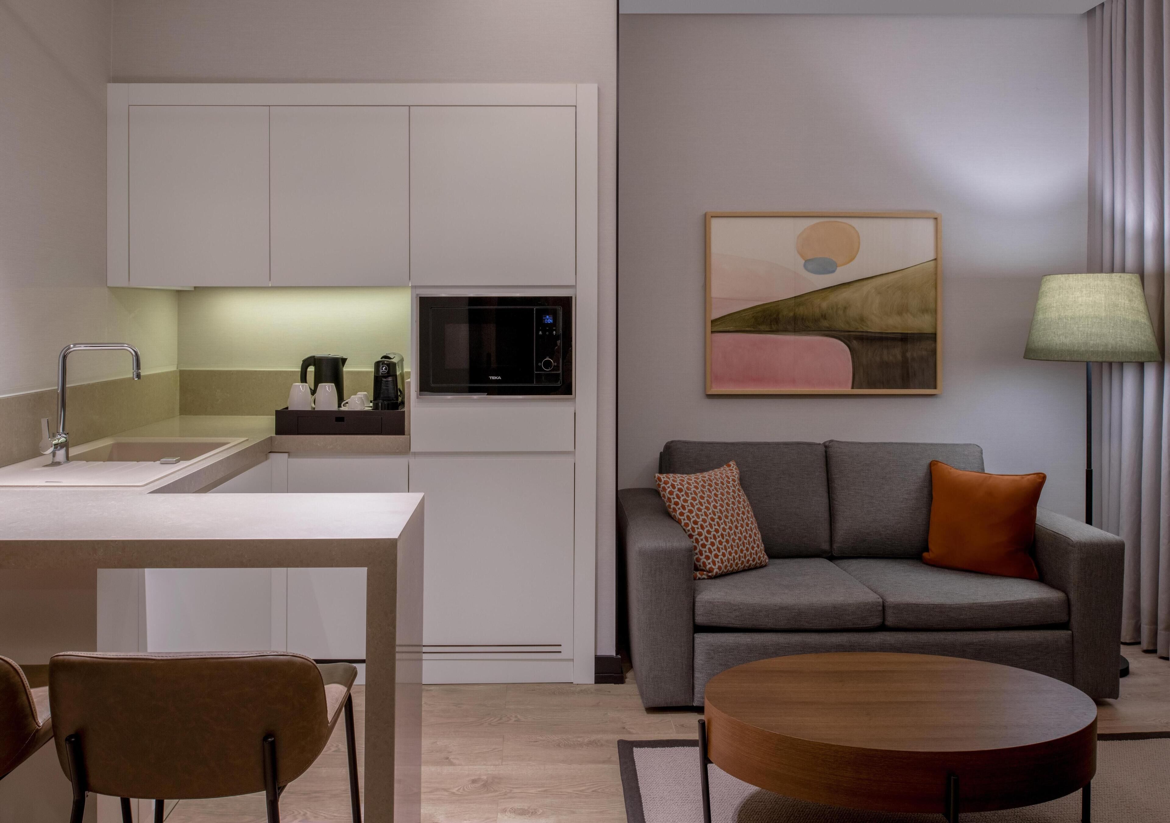 A modern kitchen with built-in appliances, a dining area with a table and chairs, a gray sofa with cushions, and a painting on the wall.