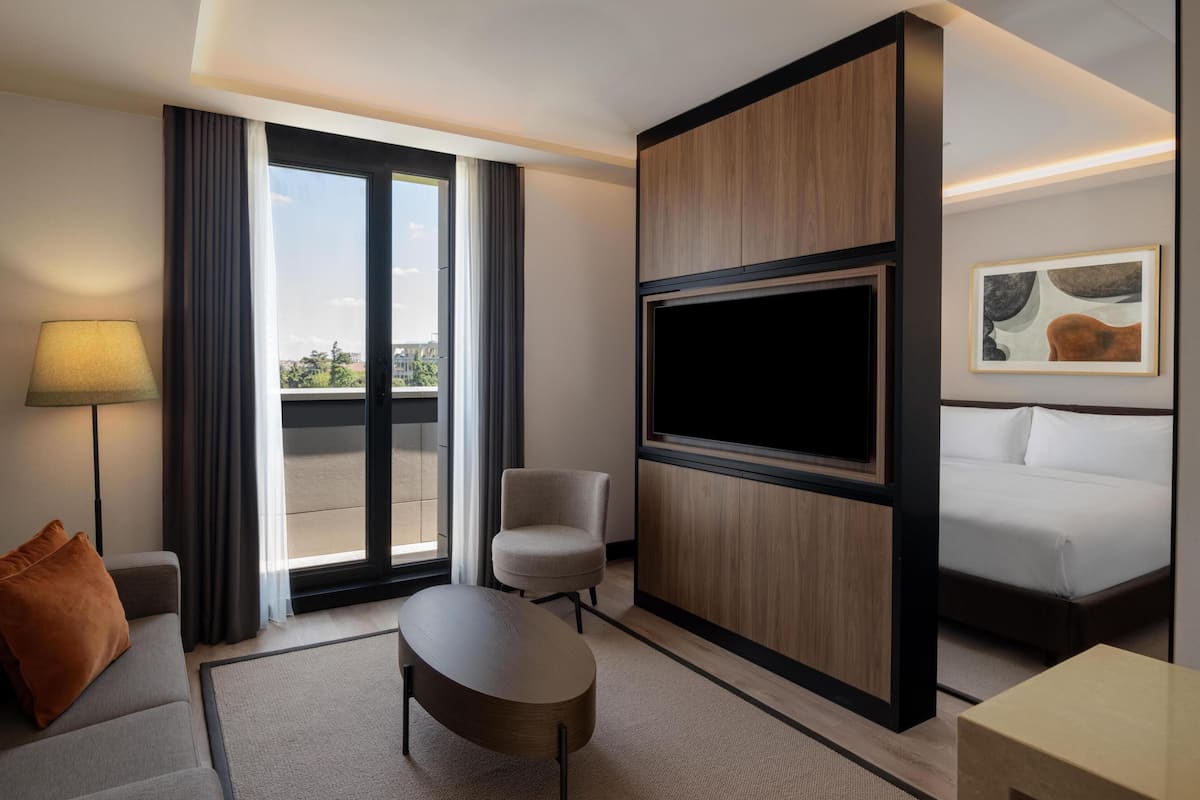 A hotel room with a sofa, armchair, TV, bed, and a view through the window.