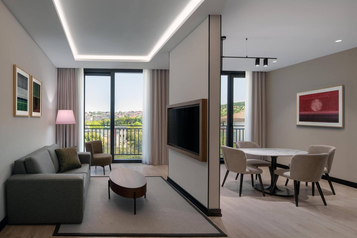A modern hotel room with a sofa, a dining table with chairs, a TV, and a balcony with a city view.