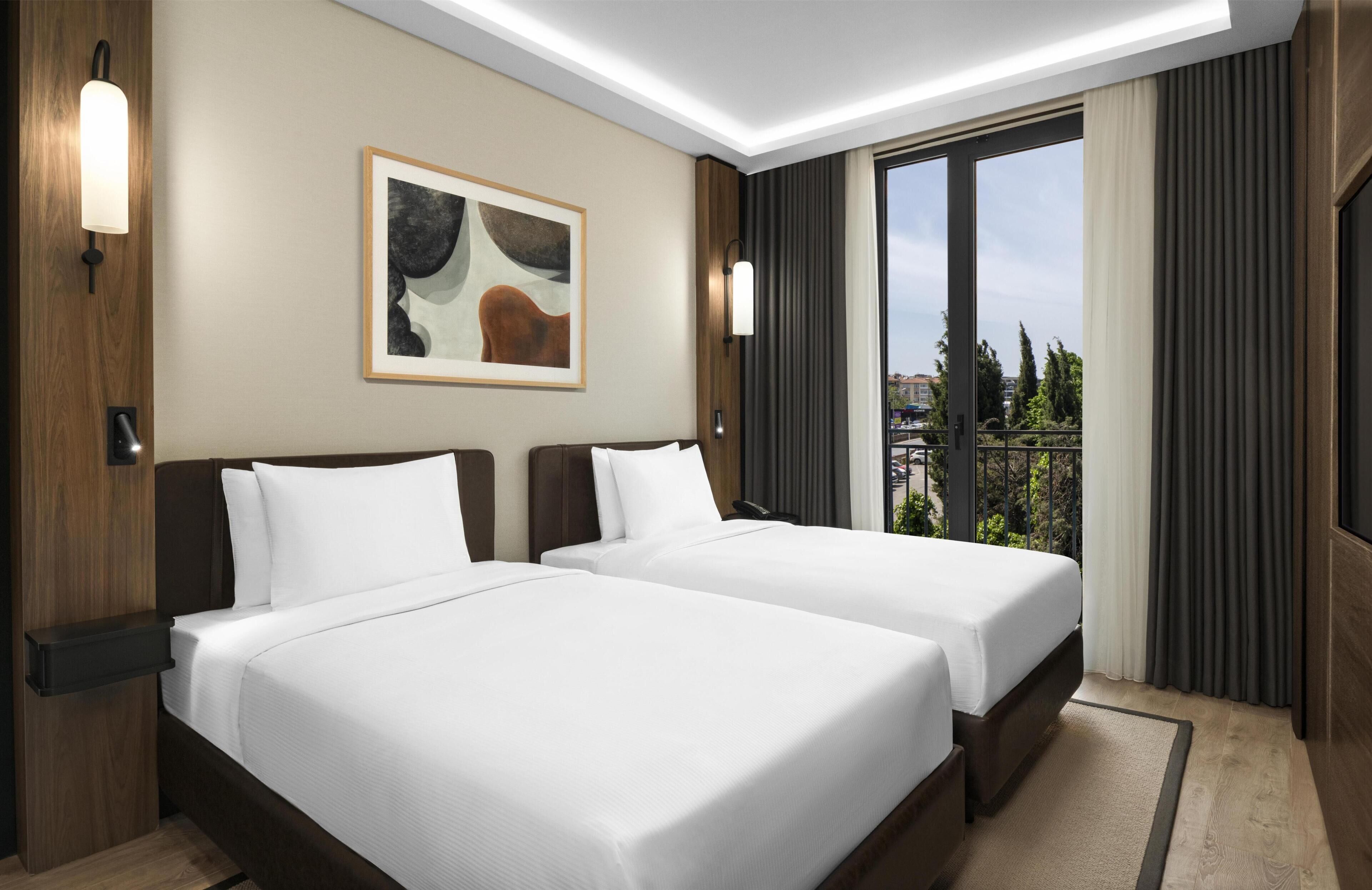 A hotel room with two beds, a painting on the wall, and a view through the window.