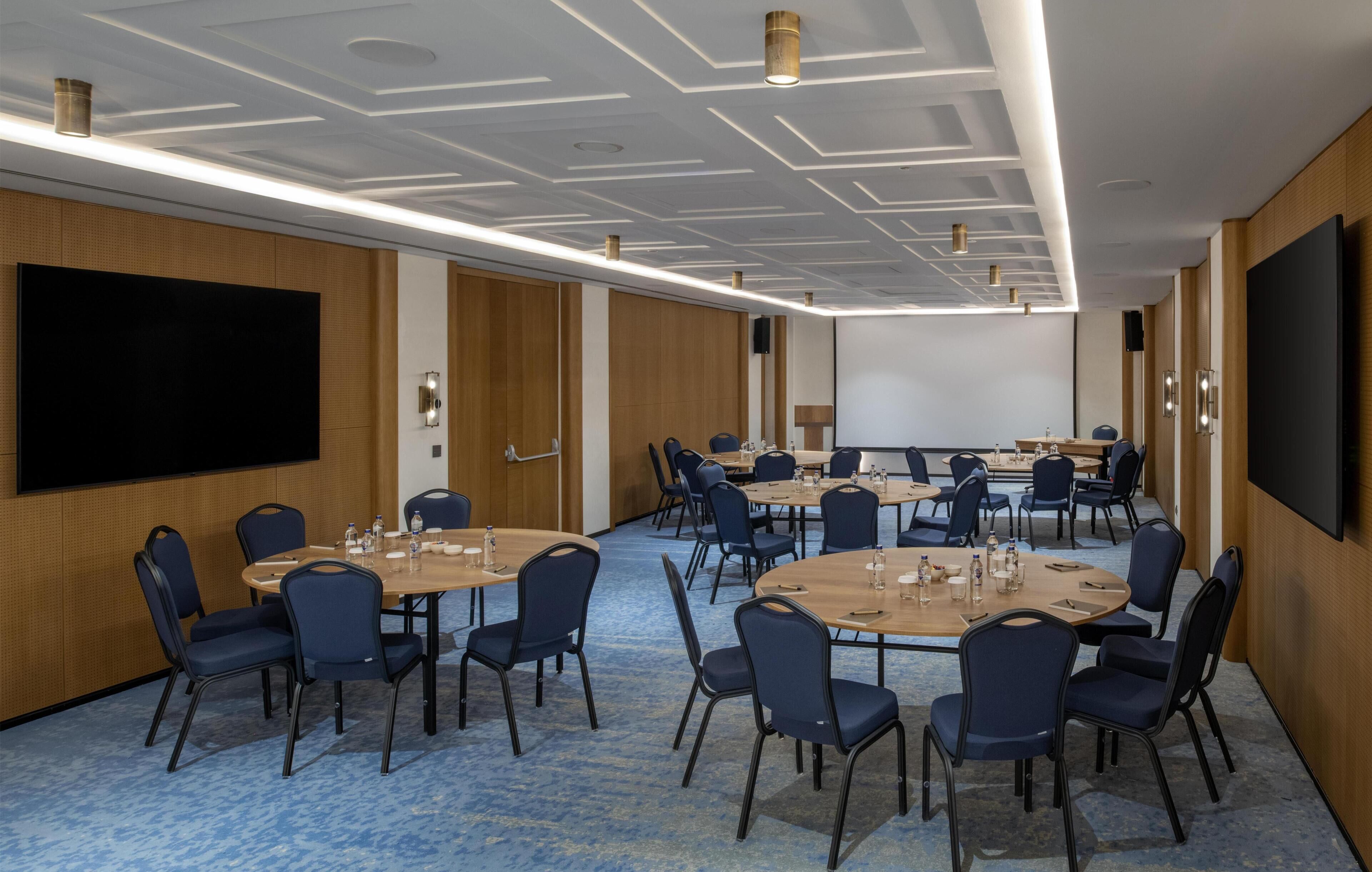A conference room with multiple tables and chairs, a large screen, and wooden paneling on the walls.