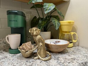 Eine Küchenzeile mit einer grünen Keurig-Kaffeemaschine, einer weißen Tasse, einer goldenen Affigungsfigur, einem gelben Kaffeekocher und einer Holztasse.