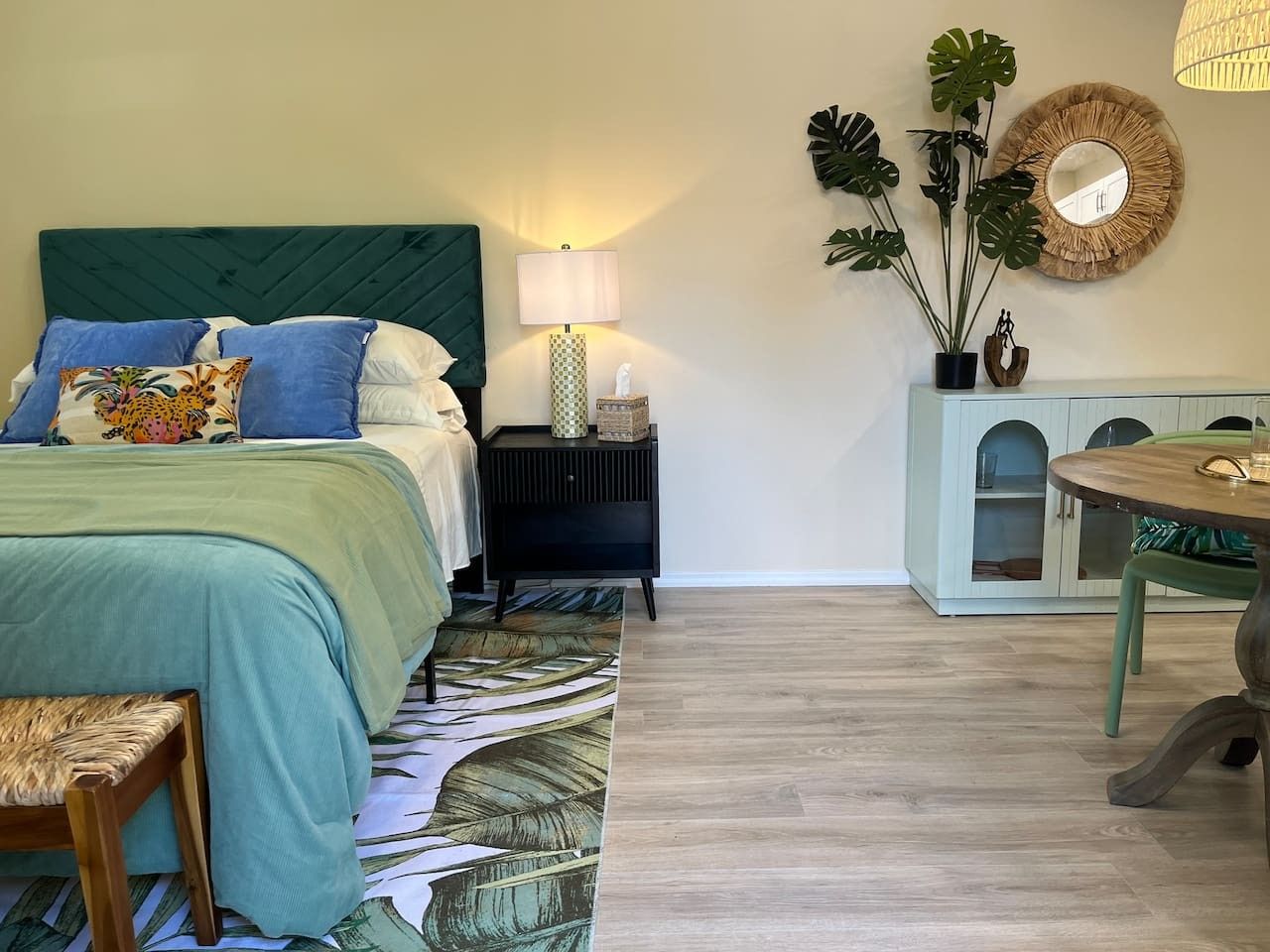 A bedroom with a bed, bedside table, lamp, potted plants, and a small cabinet.