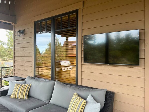 A covered outdoor seating area with a gray sofa and cushions, a flat-screen TV mounted on the wall, and a grill on the adjacent wooden deck.