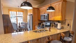Una cocina con encimeras de granito, armarios de madera, un refrigerador de acero inoxidable, un microondas y una estufa. Hay taburetes de bar y una mesa de comedor con sillas.