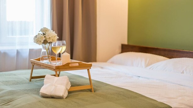 Una cama bien hecha con una bandeja que sostiene una copa de vino blanco, un florero con flores blancas y una toalla doblada sobre un edredón verde.