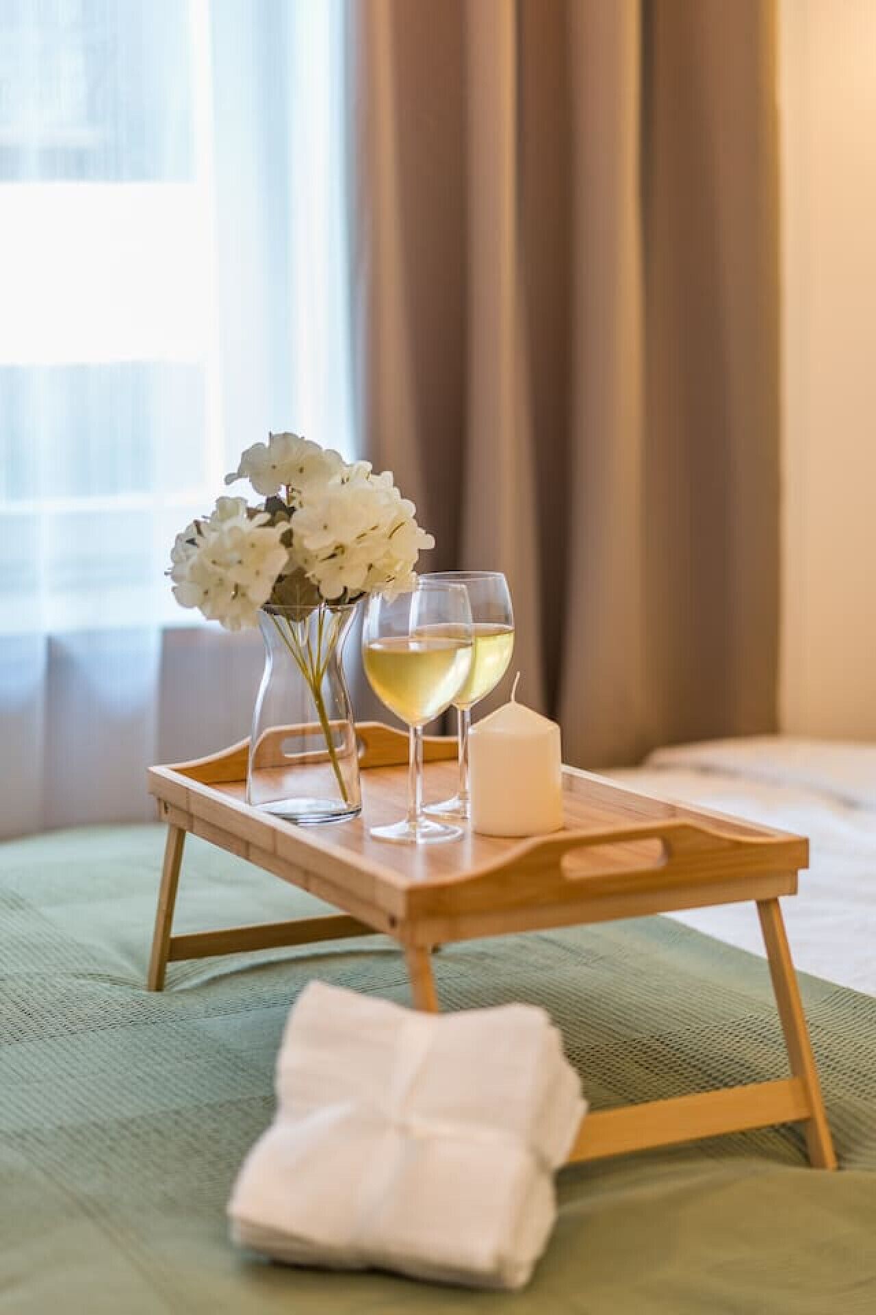A tray with two glasses of white wine, a candle, and white flowers on a bed.