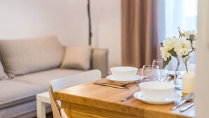 Un área de comedor con una mesa de madera dispuesta para dos personas, un sofá al fondo y un florero con flores blancas sobre la mesa.