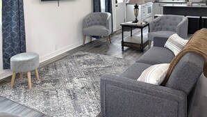 Un salon moderne avec un téléviseur à écran plat, des canapés gris, un tapis à motifs et un ventilateur de plafond.