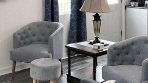 Un salon moderne avec des fauteuils à revêtement gris, une petite table avec une lampe et une vue sur une cuisine à travers une fenêtre ornée de rideaux à motifs.