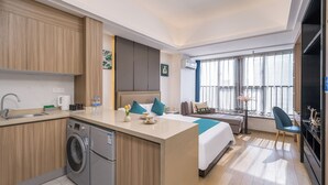 A modern hotel room with a kitchen area, a bed with blue pillows, a seating area with a table and chairs, and a large window with curtains. - Guangzhou Hantao International Apartment (Guangzhou)