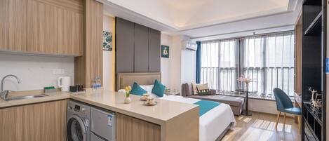 Una habitación de hotel moderna con cocina, una cama con almohadas azules, una zona de estar con mesa y sillas, y un ventanal con cortinas.