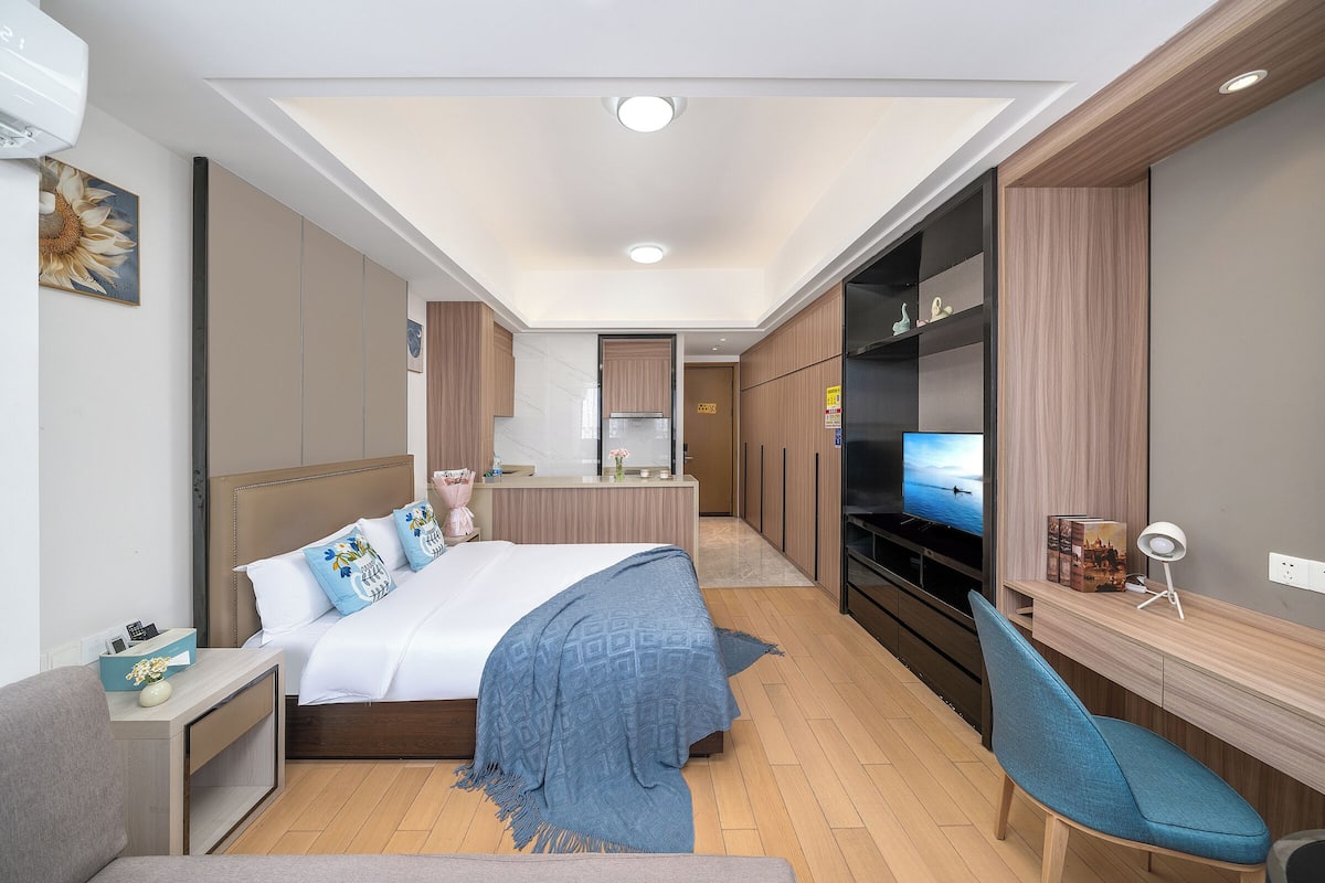 A modern hotel room with a bed, bedside table, TV, and a desk with a chair.