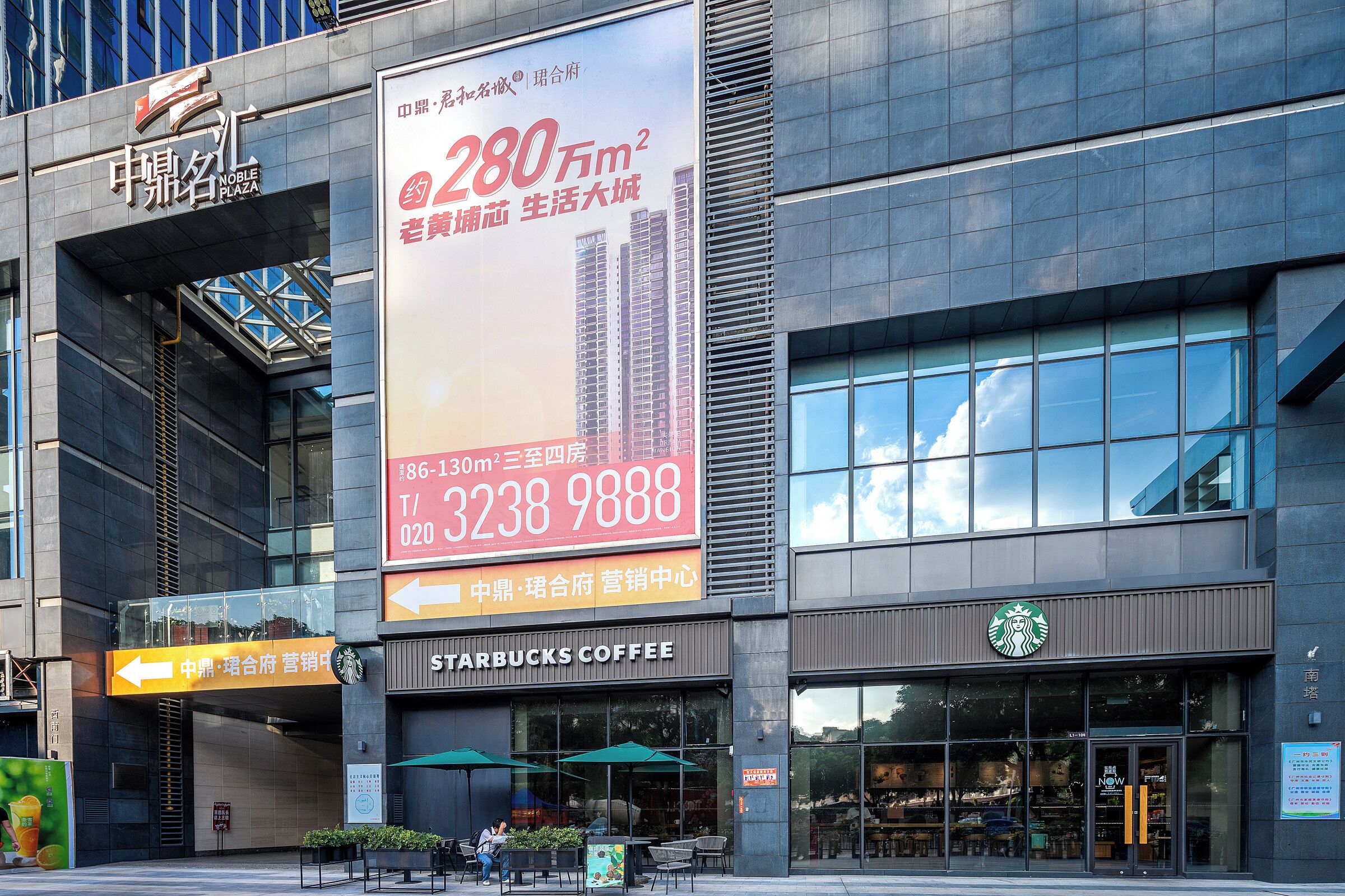 A modern building with a Starbucks Coffee shop, a large billboard advertising a property, and a sign indicating the direction to the mall.