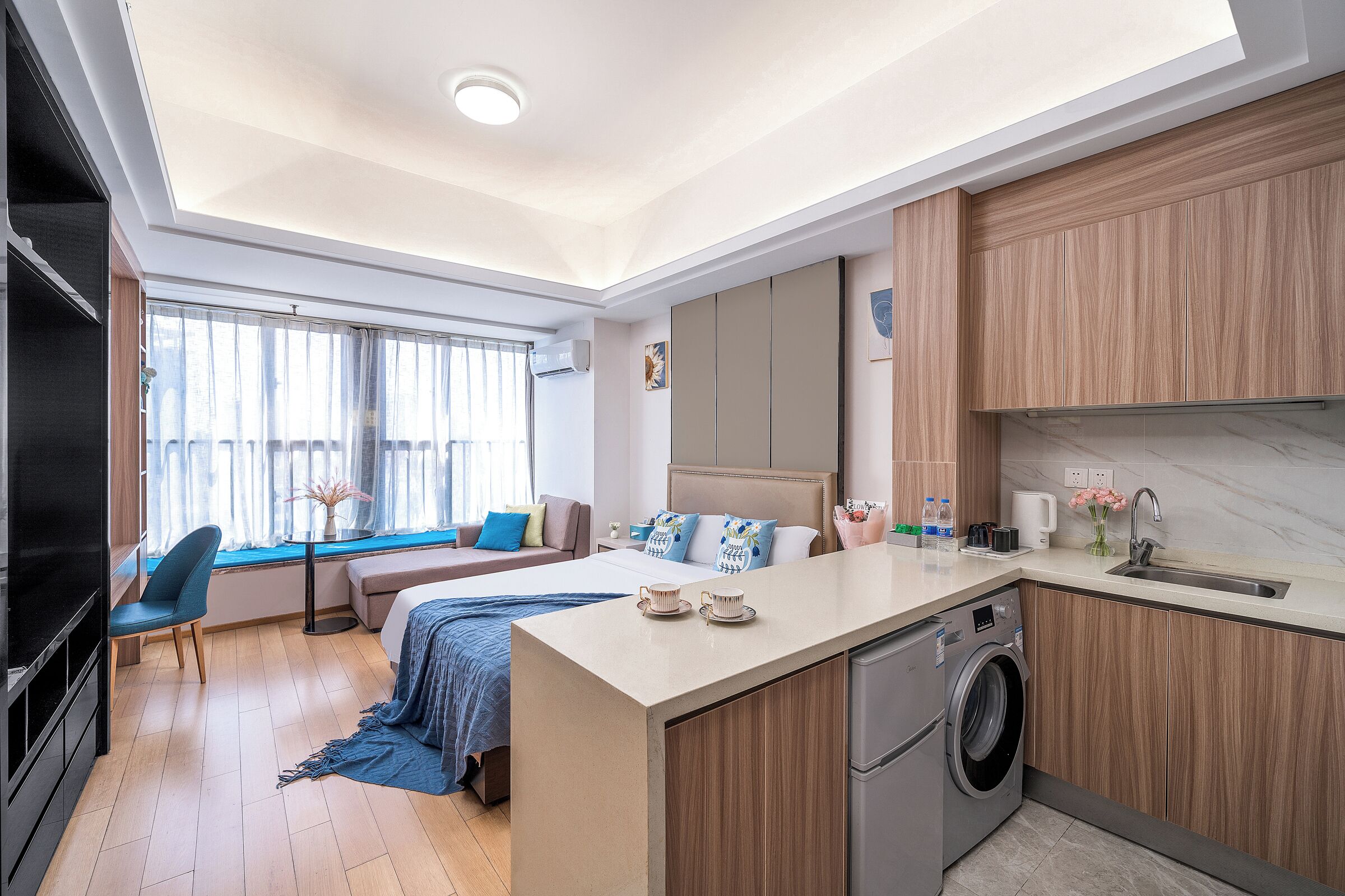A modern hotel room with a large bed, a sitting area, a kitchenette, and a washing machine.