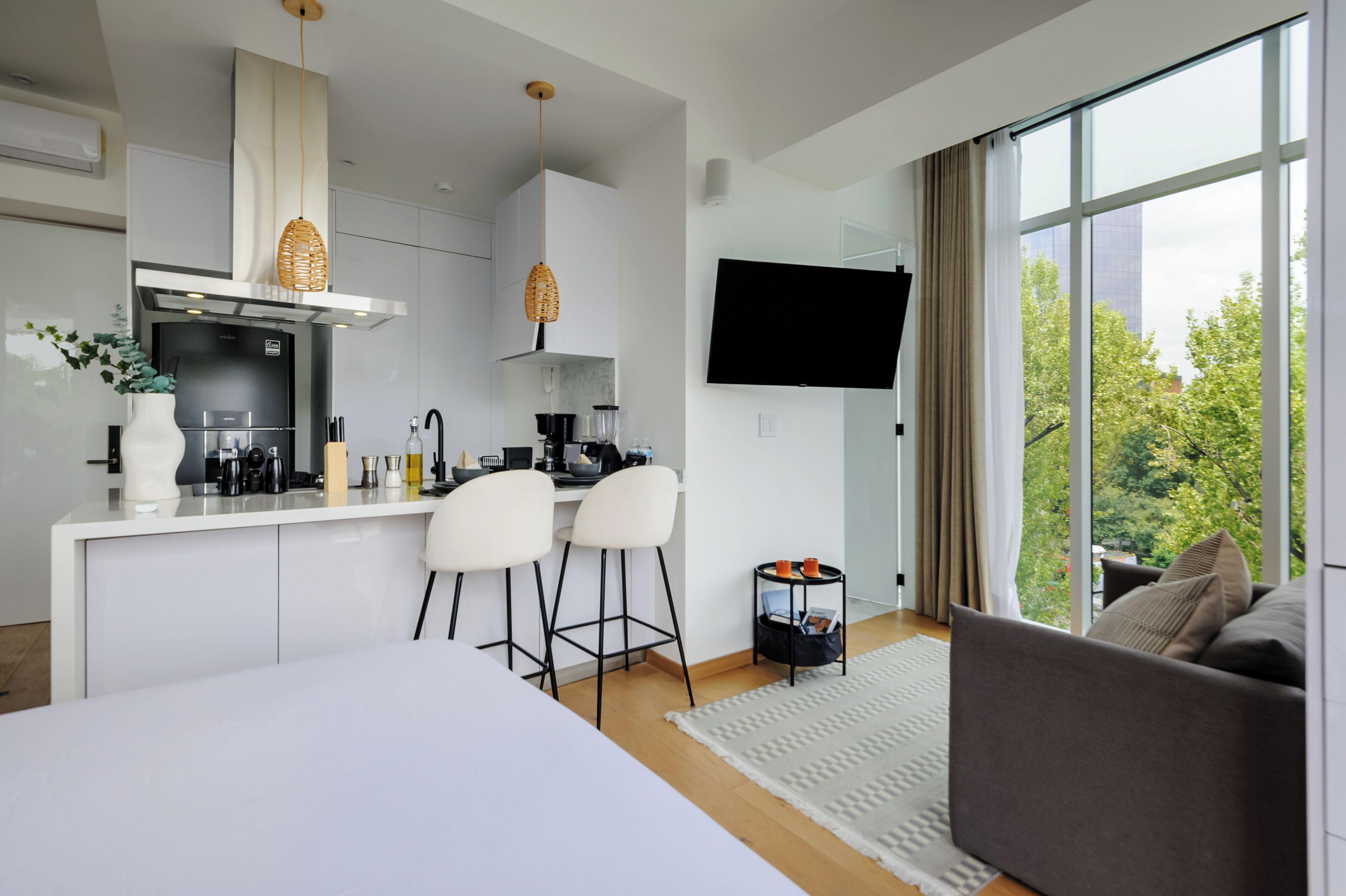 A modern kitchen with white cabinets, a dining area with two white chairs, a sofa, and a TV mounted on the wall.