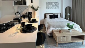 Ein modernes Hotelzimmer mit Küchenzeile, einem Bett mit Kopfteil, einem Nachttisch und einem großen Fenster mit Vorhängen.